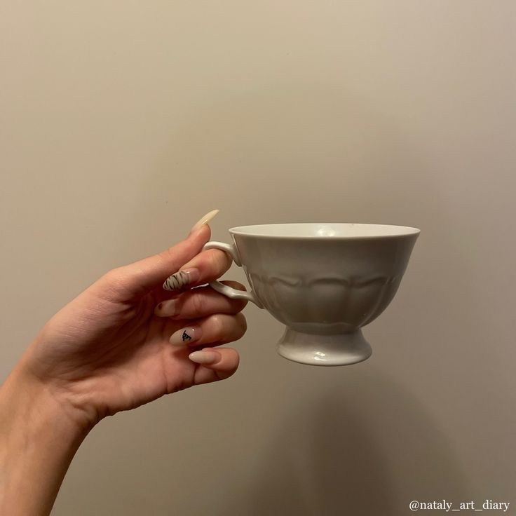 a woman's hand holding a white coffee cup in front of a beige wall