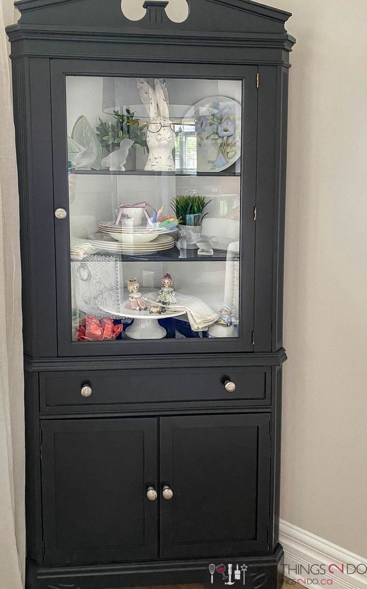 a black china cabinet with glass doors and shelves on the top, in front of a white wall