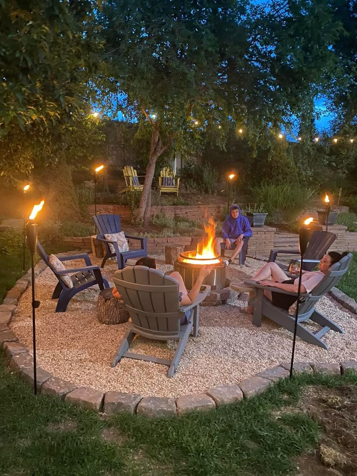 an outdoor fire pit surrounded by lawn chairs and lights with people sitting around it at night