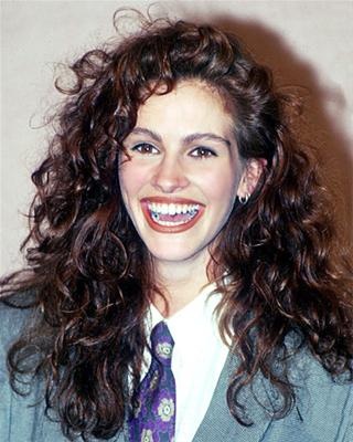 a woman with long curly hair wearing a suit and smiling at the camera while standing in front of a white wall