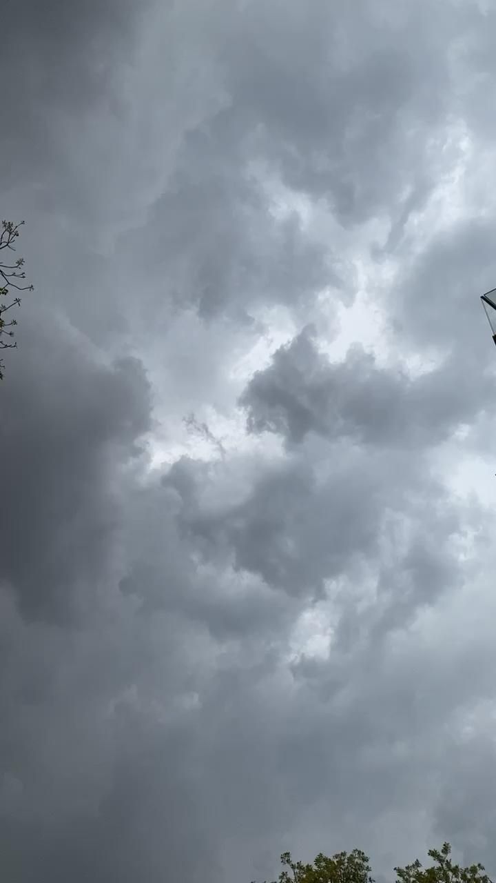 an airplane flying in the cloudy sky with trees around it and dark clouds above them