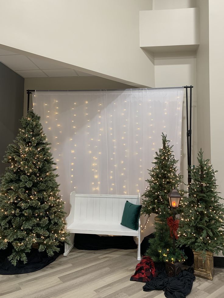 three christmas trees sitting in front of a white wall with lights on it and a bench next to them