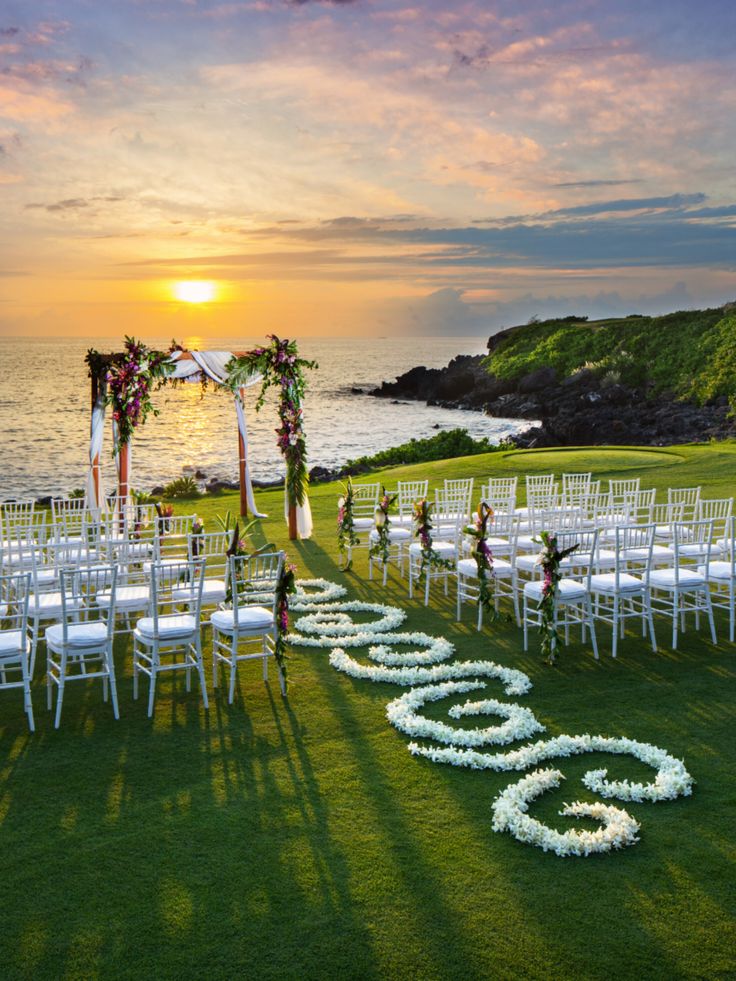 an outdoor wedding setup with white chairs and flowers on the grass near the ocean at sunset