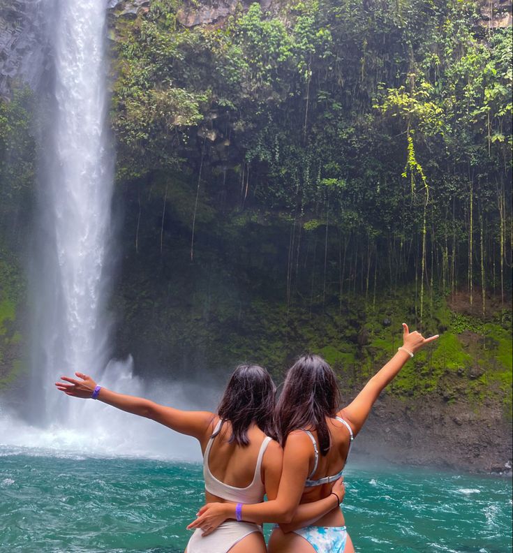 two women in bikinis standing next to a waterfall and holding their arms out towards the water