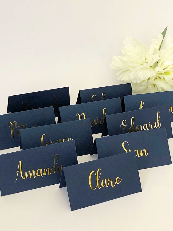 blue place cards with gold lettering on them and a white flower in the middle, sitting next to each other