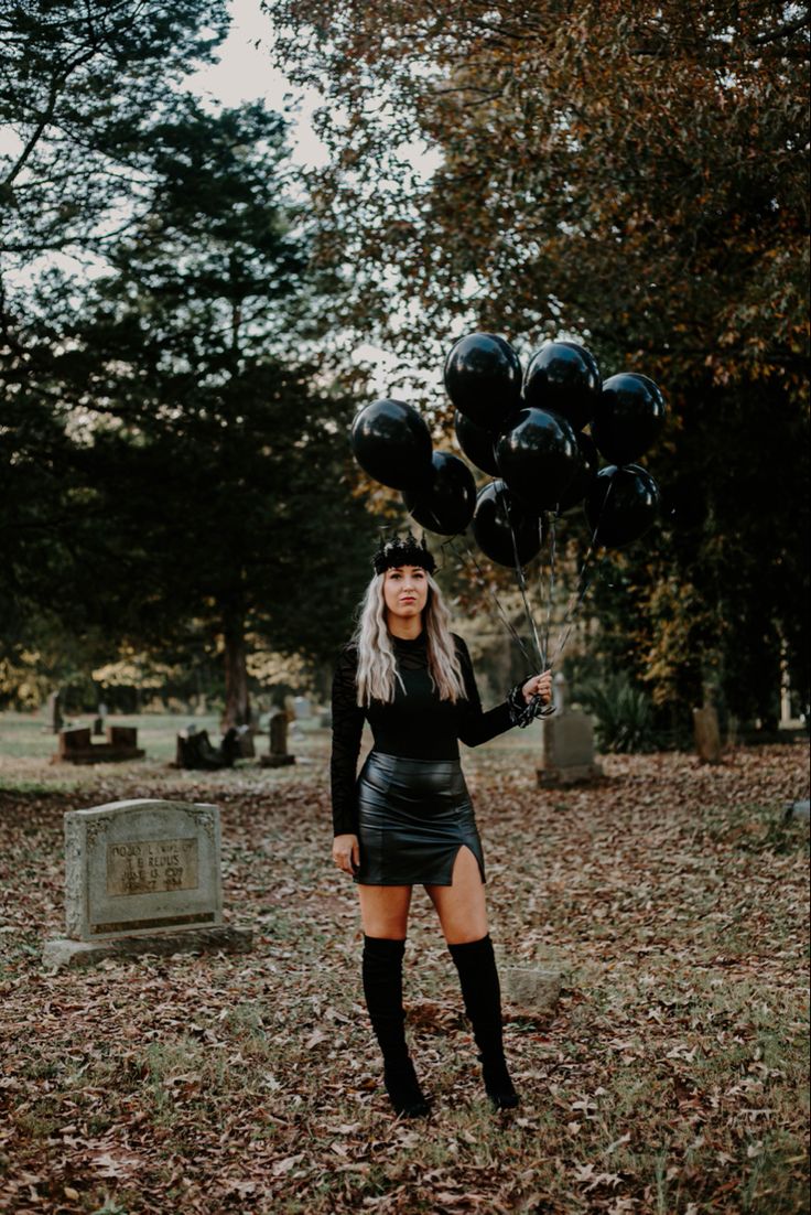 a woman holding black balloons while standing in the grass with her legs crossed and wearing knee high boots