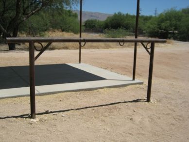 an empty swing set in the middle of a dirt field with trees and bushes behind it