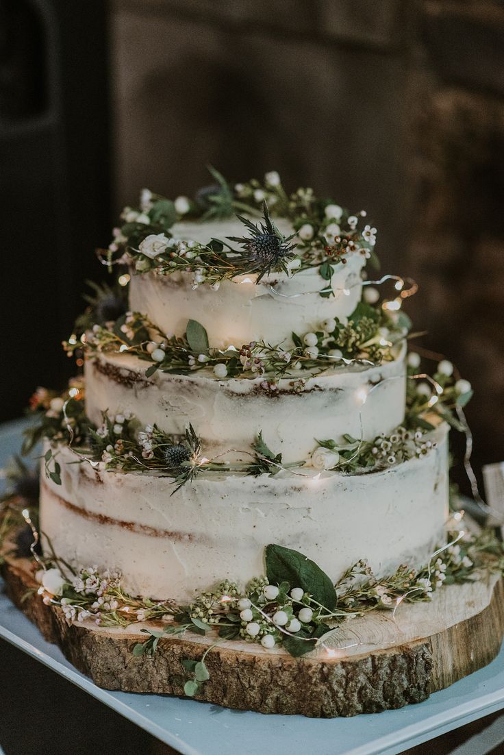 a three tiered wedding cake with greenery and lights on the top is sitting on a table