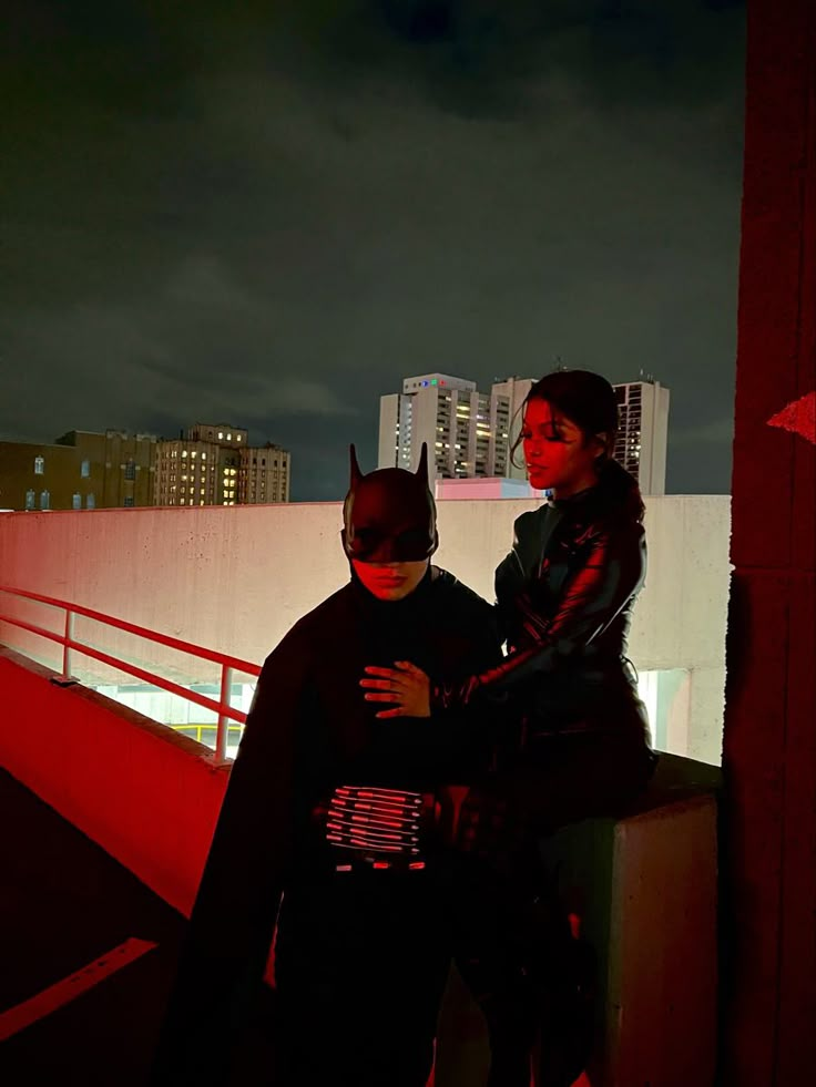 two people dressed up as batman and catwoman standing on a bridge at night with city lights in the background