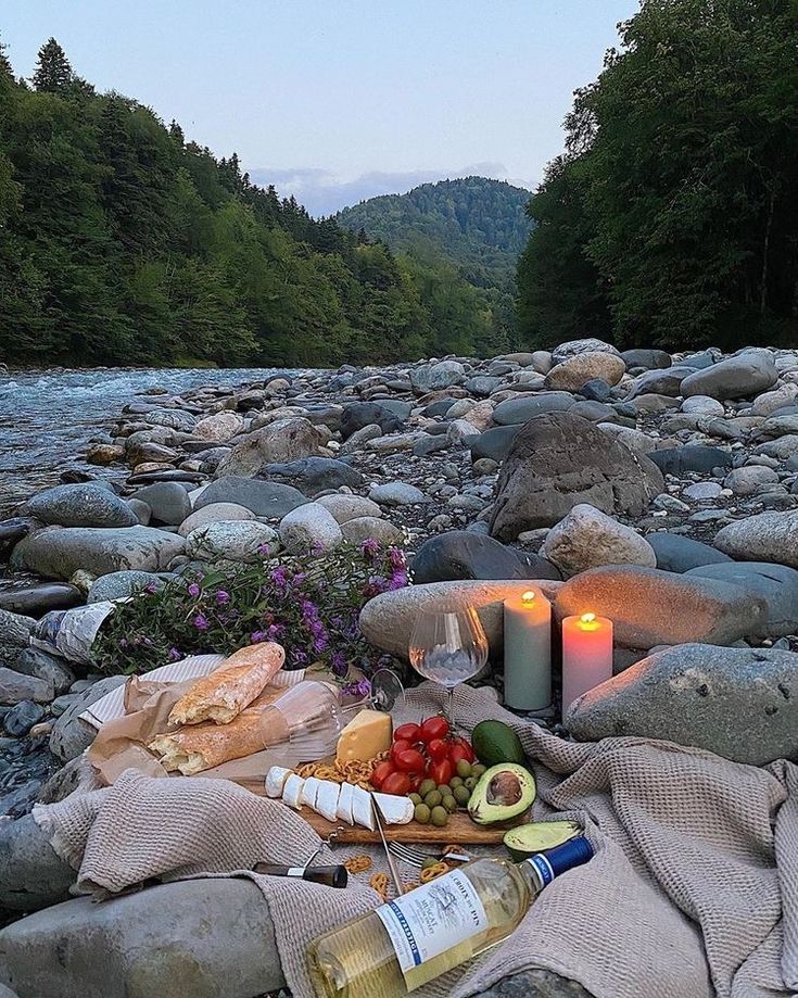 food and drinks are laid out on the rocks near a river with mountains in the background