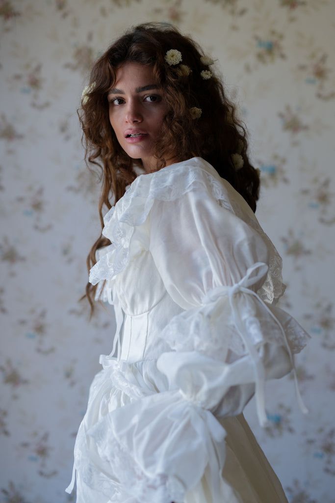 a woman in a white dress with flowers on her head and long curly hair standing against a floral wallpapered background