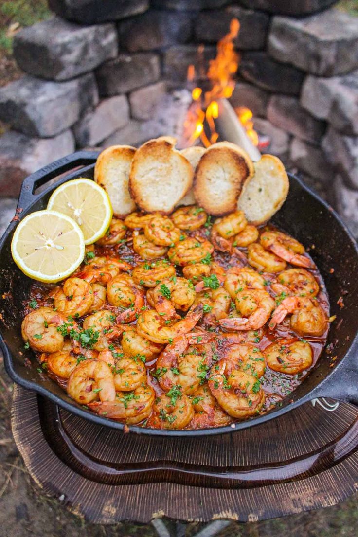 shrimp and bread cooking in a skillet over an open fire with lemon wedges