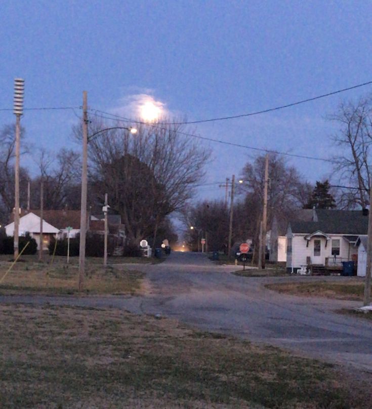 an empty street at night with the moon in the sky and houses on the other side