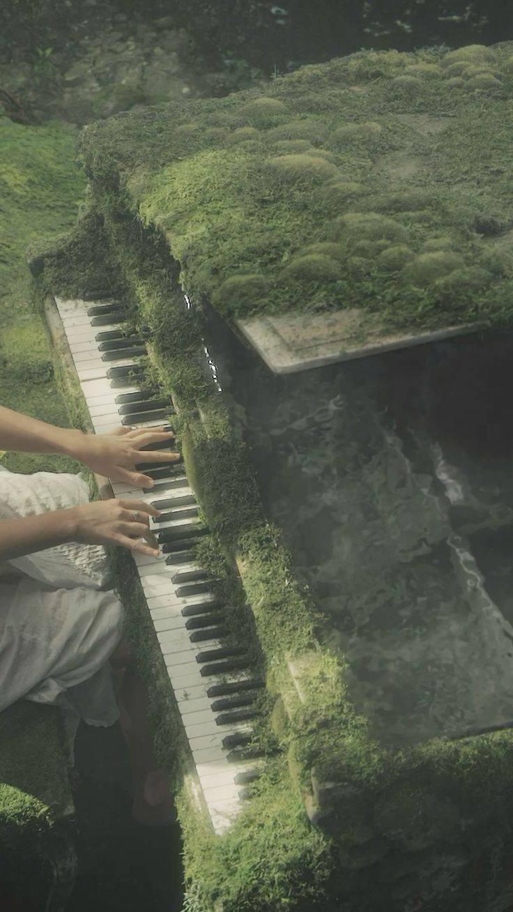 a woman sitting on top of a lush green field next to a giant piano covered in moss