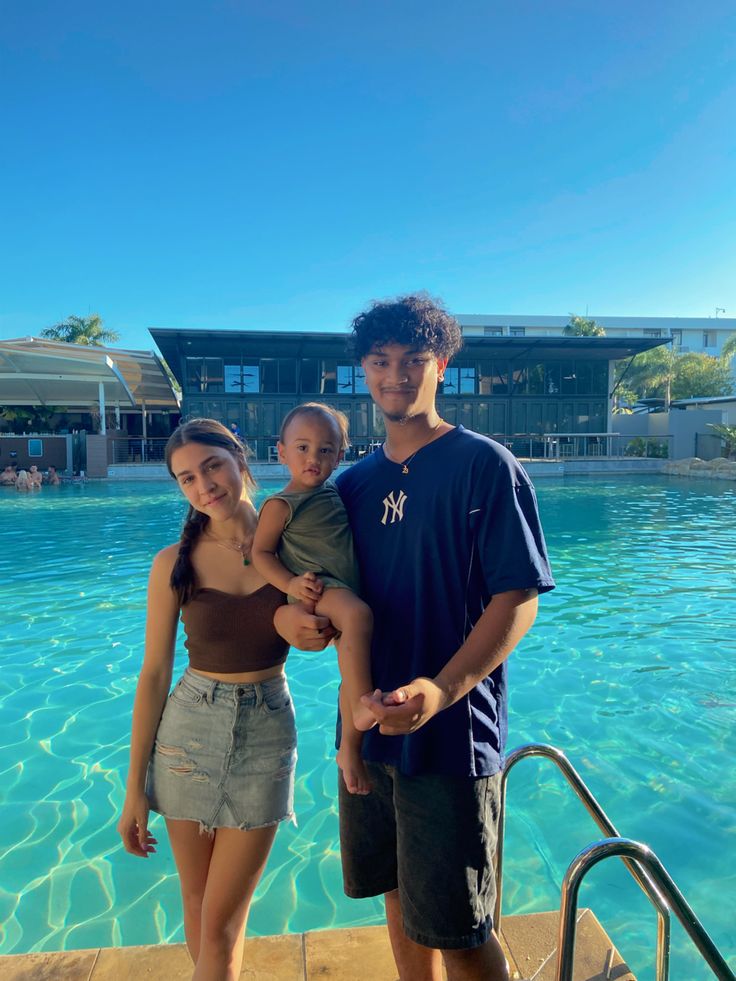 a man and woman standing next to a swimming pool holding a small child in their arms