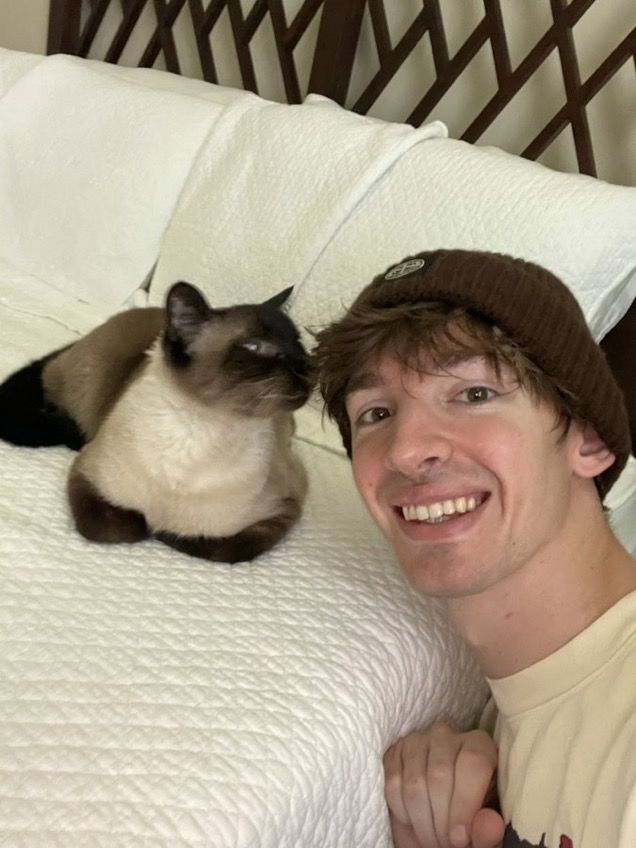 a man laying on top of a bed next to a siamese cat in a hat