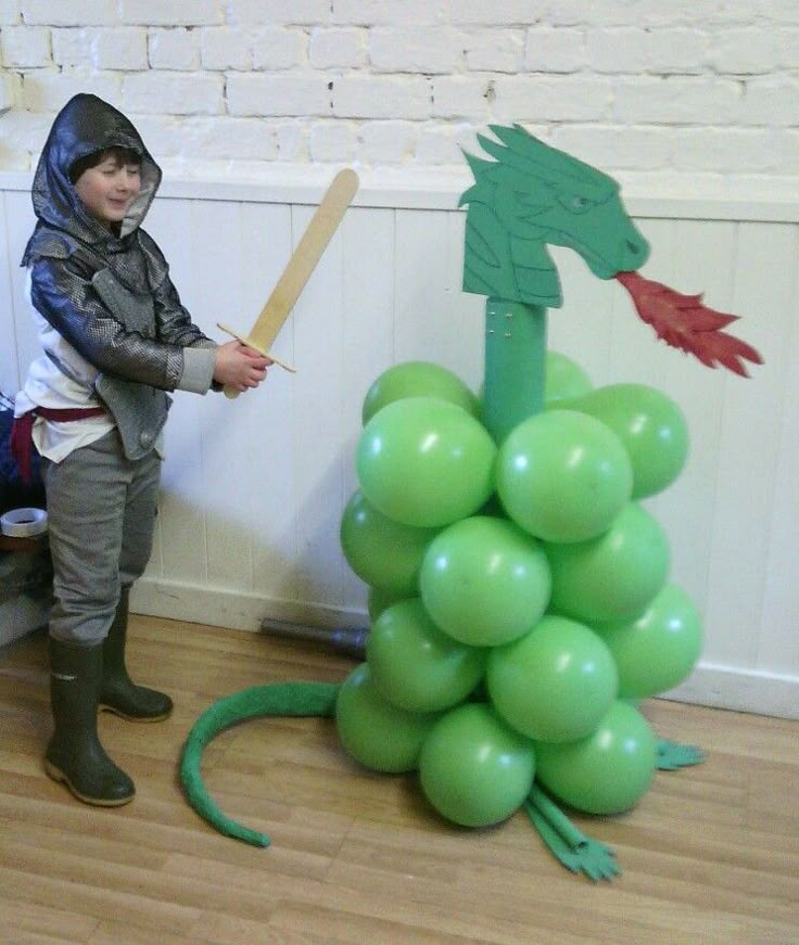 a young boy dressed as a dragon holding a baseball bat next to a bunch of green balloons