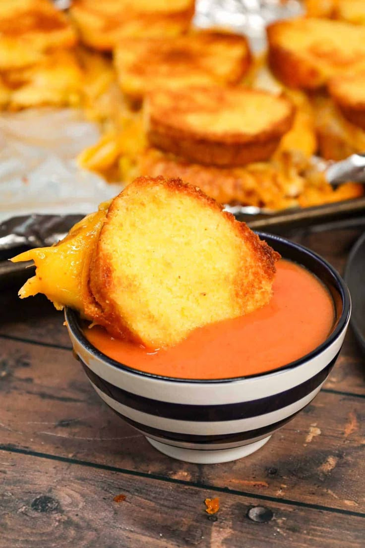 a piece of bread sticking out of a bowl of tomato soup with other food in the background