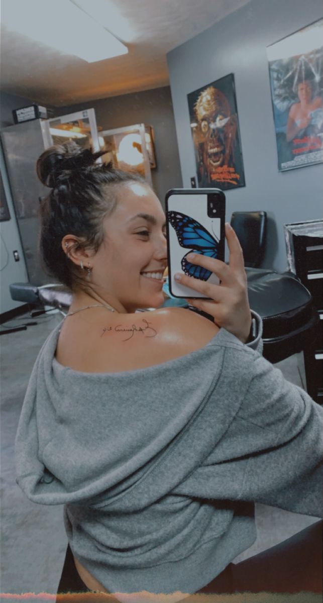 a woman taking a selfie with her cell phone in an office setting, while wearing a butterfly tattoo on her shoulder