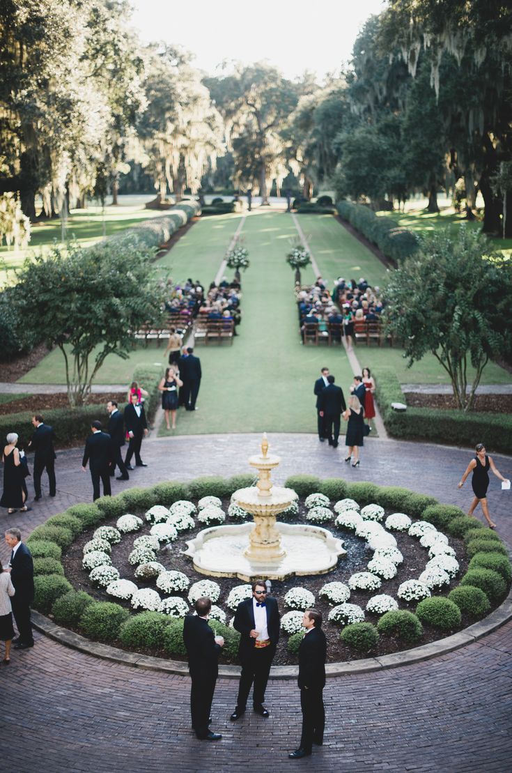 two men standing in front of a fountain surrounded by flowers and greenery with people walking around