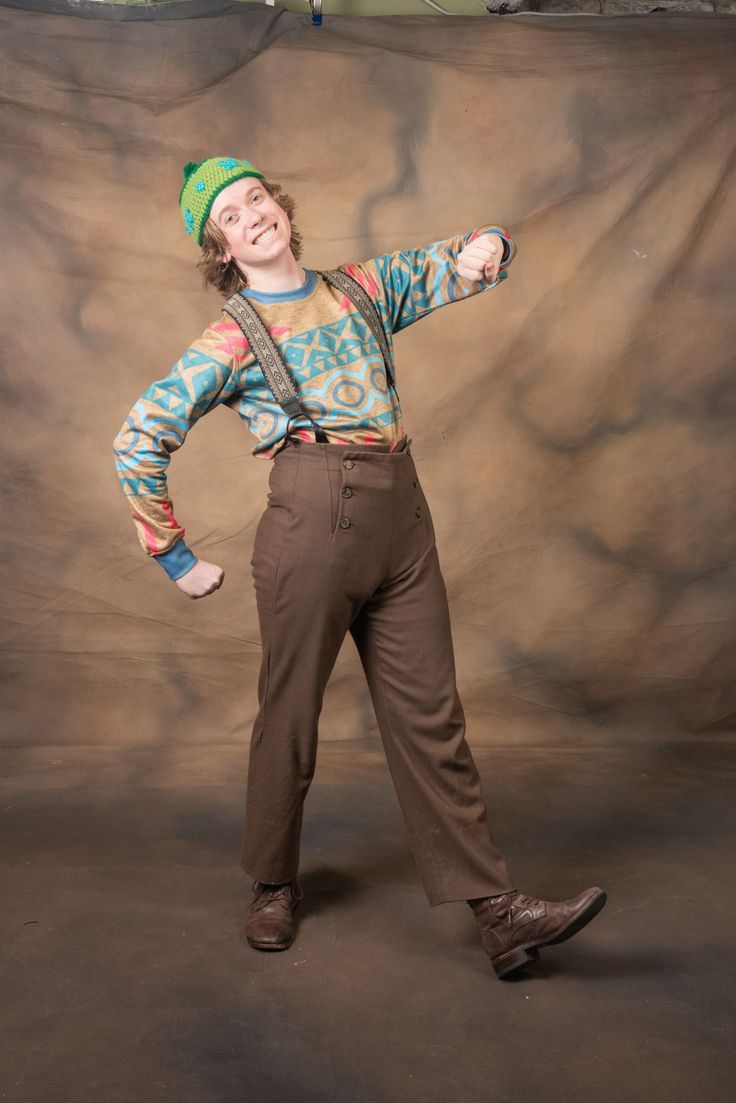 a young man standing in front of a wall wearing brown pants and a green hat