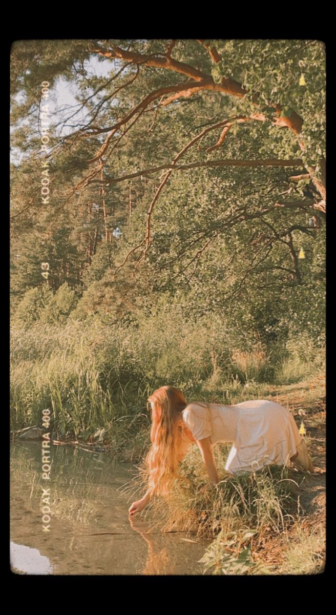 a woman sitting on the bank of a river with her head down and long hair blowing in the wind