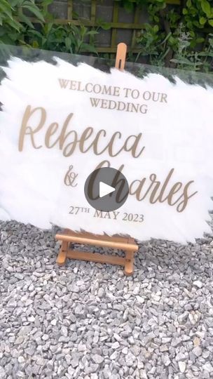 a welcome sign for a wedding with white feathers and gold lettering on it, sitting in gravel