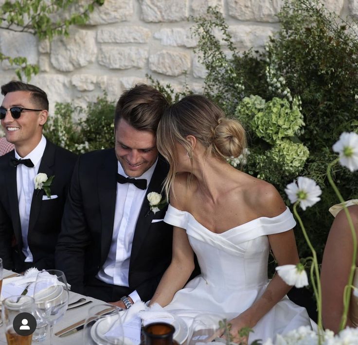 two men and a woman in formal wear sitting at a table with flowers on it