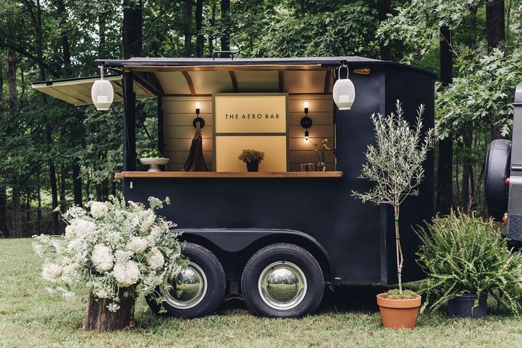 a black food truck parked in the middle of a forest with potted plants and trees