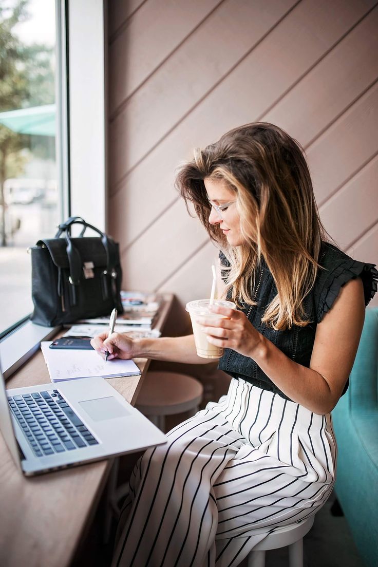 a woman sitting at a table with a laptop and coffee in front of her, writing on a piece of paper