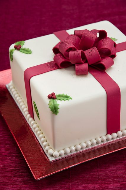 a square cake decorated with holly leaves and a red ribbon on a platter against a purple background