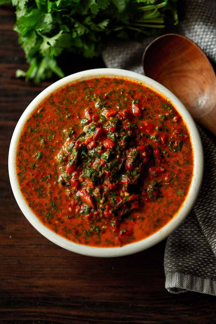 a white bowl filled with red sauce and garnished with parsley next to a wooden spoon