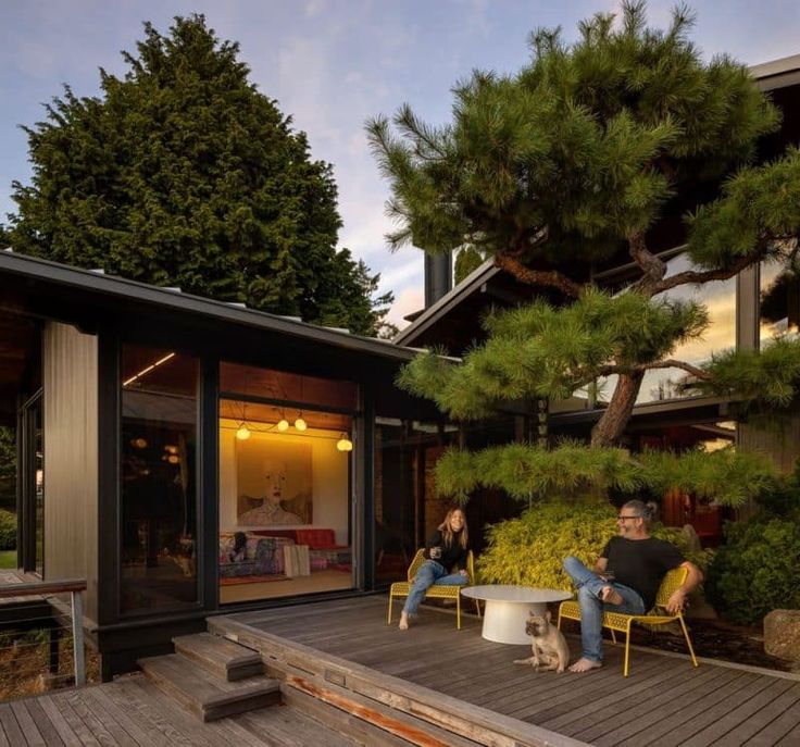 two people sitting on chairs in front of a house with pine trees and bushes behind them