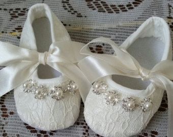 pair of white baby shoes with bows and pearls on the bottom, sitting on a lace tablecloth