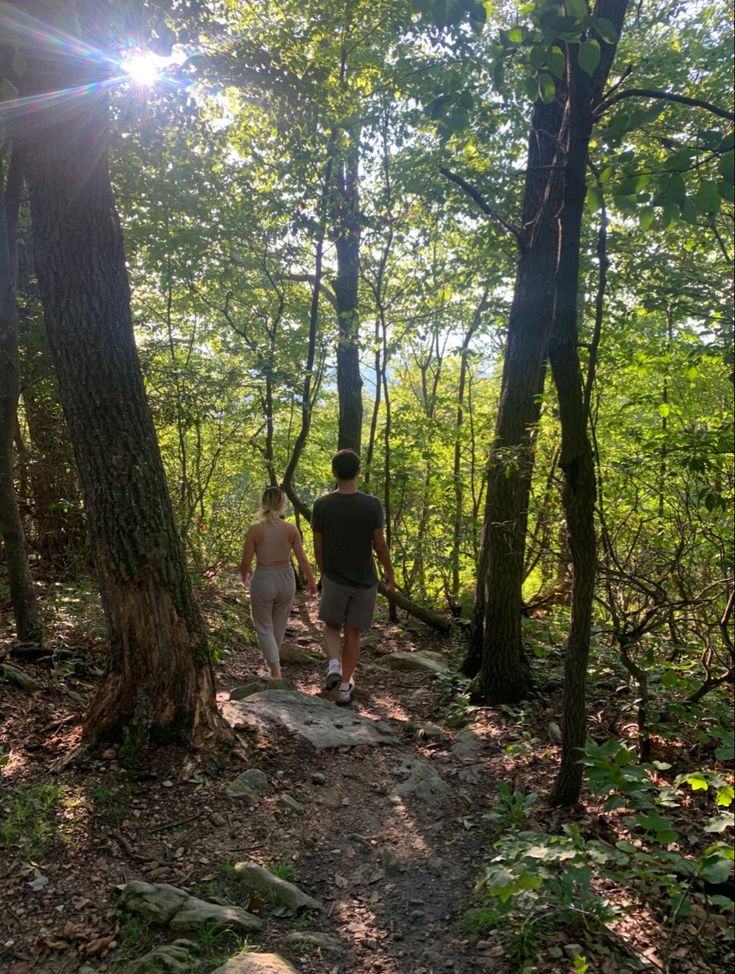 two people walking down a trail in the woods with trees and rocks on either side