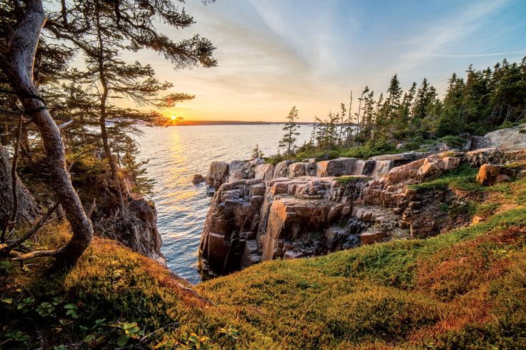 the sun is setting over an ocean with rocky cliffs and trees on either side, as seen from across the water