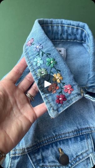 a hand holding up a jean jacket with embroidered flowers on the front and back pockets