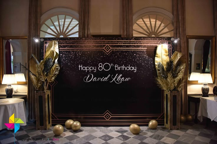 a black and gold birthday backdrop with balloons on the floor in front of two tables