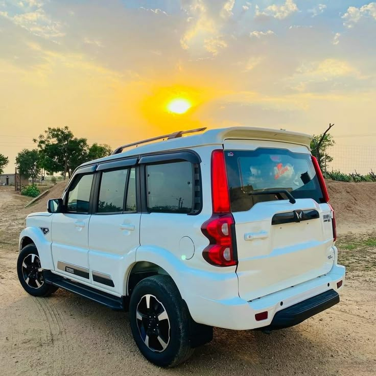 the back end of a white suv parked on top of a dirt field at sunset