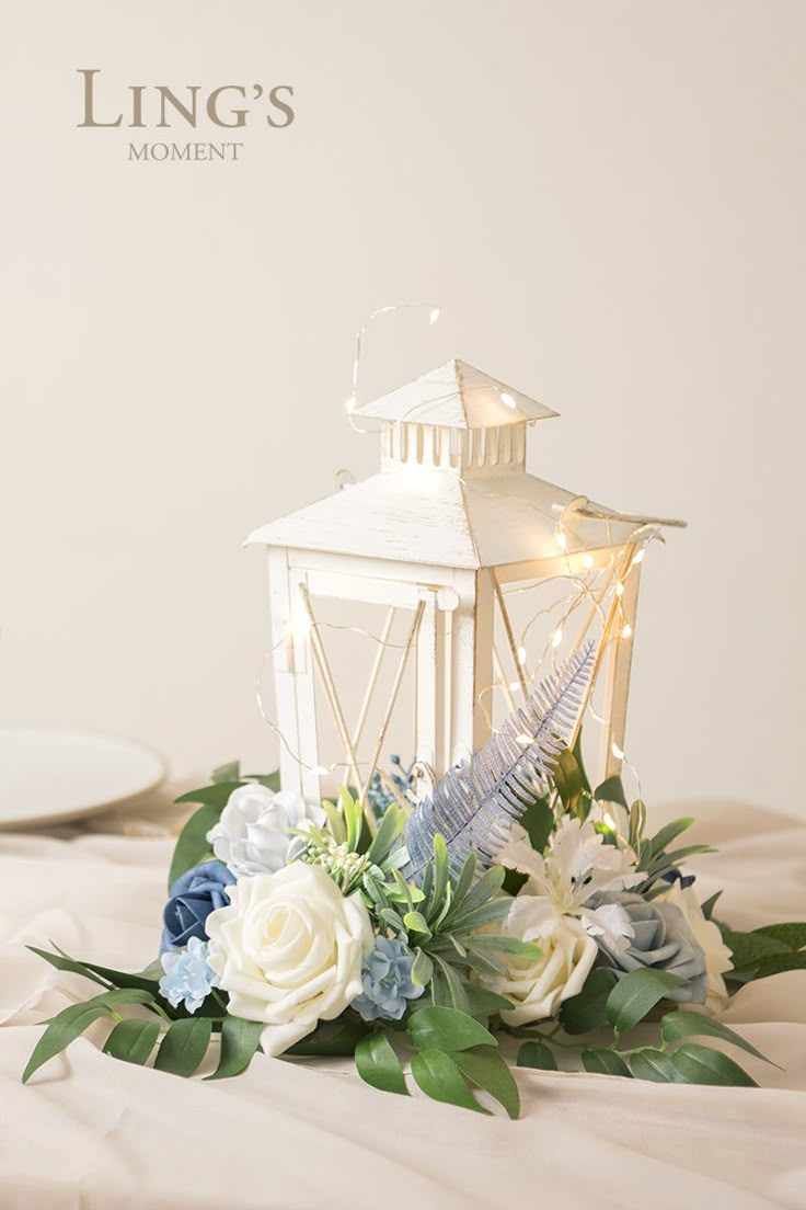 a white lantern with blue flowers and greenery on the table in front of it