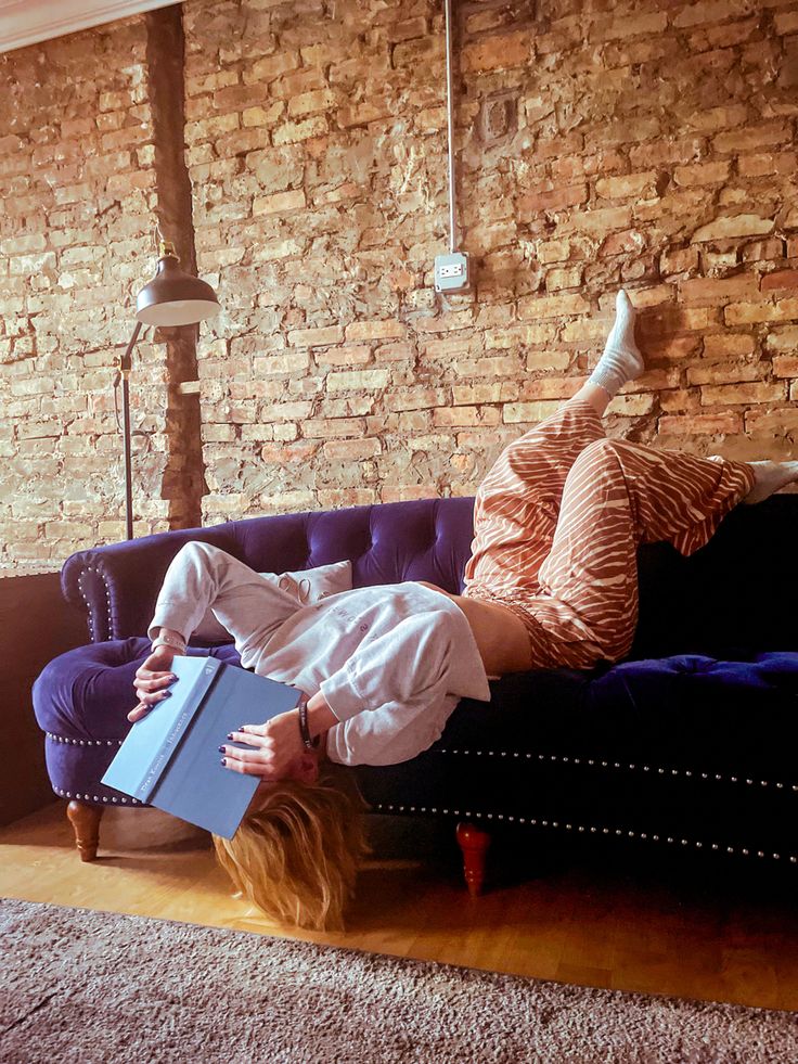 a woman laying on top of a blue couch in front of a brick wall holding a book