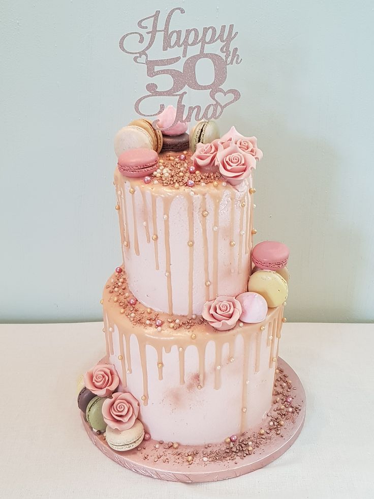 a three tiered cake with pink icing and flowers on top is decorated with macaroons