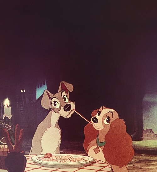 lady and the tramp sitting at a dinner table eating spaghetti from a plate in front of them