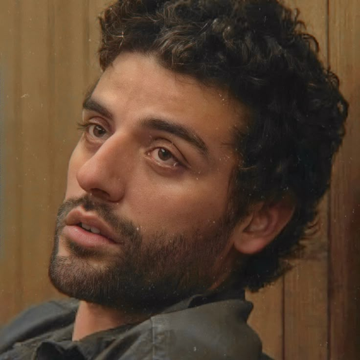 a man with curly hair and beard stares into the distance while standing in front of a wood paneled wall