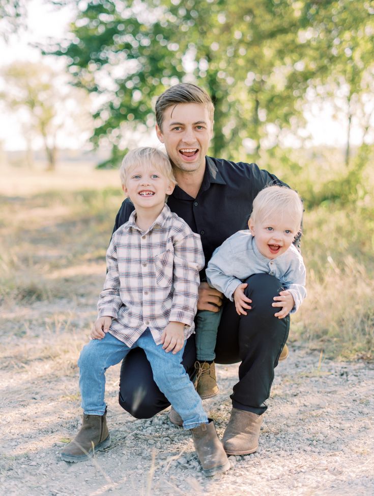 a man kneeling down with two young boys on his lap and smiling at the camera