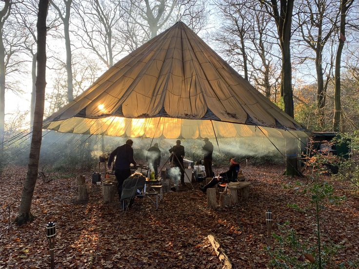 several people sitting around a campfire in the woods under a large, brown tent