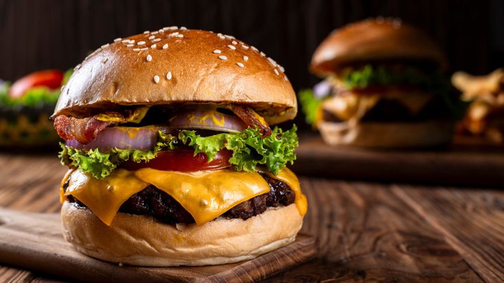 a cheeseburger with bacon and lettuce on a wooden cutting board next to other burgers