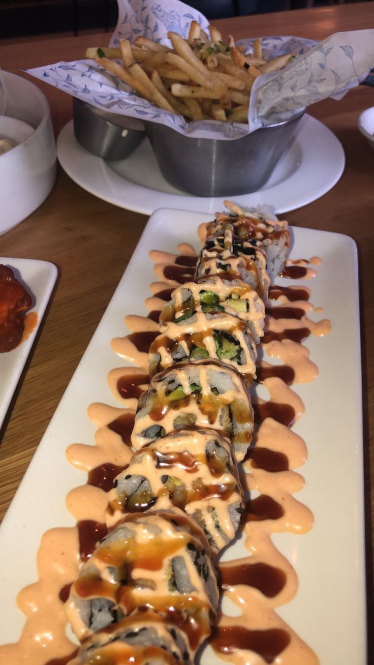 a long white plate topped with sushi covered in sauce next to fries and coleslaw