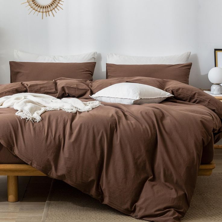 an unmade bed with brown sheets and pillows in a white walled room next to a wooden table