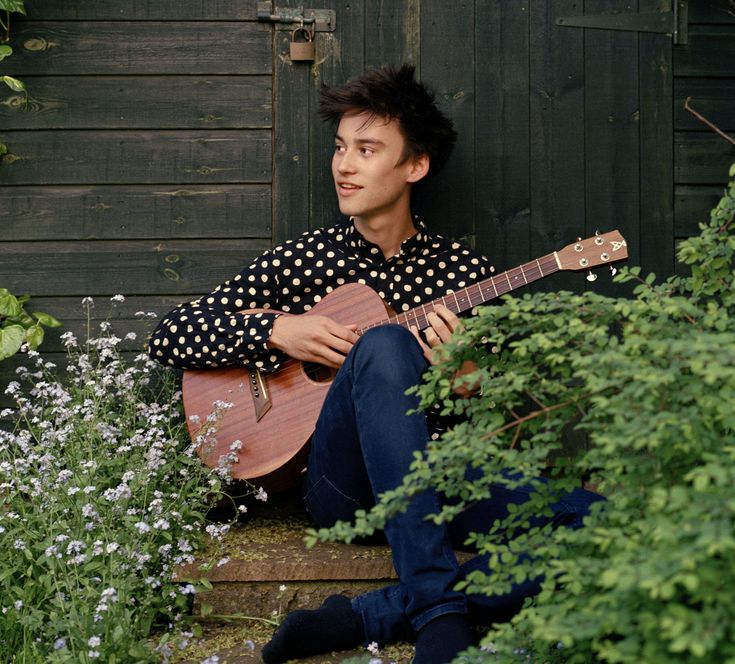 a man sitting on the ground with a guitar in his hand and plants around him